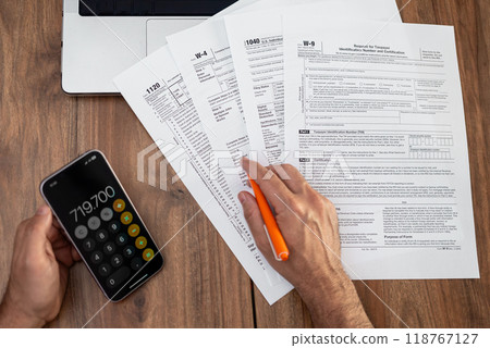 Man calculating taxes with forms, laptop, and smartphone on a wooden desk. Financial planning and tax preparation concept.  118767127