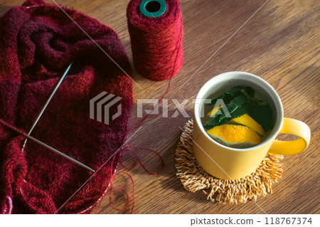 Lemon and mint leaf in yellow cup and dark red knitting with knitting needles. Autumn lifestyle. Cherry coloured wool yarn with herbal tea on the table. Knitted cardigan sweater and hot drink. 118767374