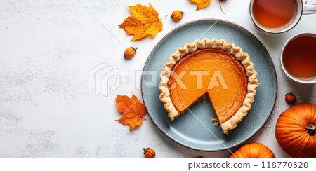 Whole pumpkin pie with a missing slice, surrounded by fall leaves, pumpkins, and two cups of tea. Cozy autumn dessert concept. 118770320