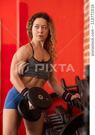 Middle aged woman takes dumbbell from rack in gym. Vertical photo. Middle aged woman takes dumbbell from rack in gym. Vertical photo. 118772016