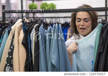 Portrait of a fat woman in a plus size store through hangers with clothes.  118772017