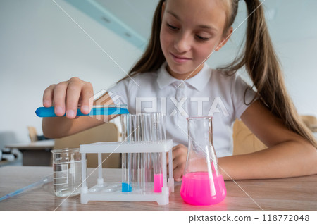 A schoolgirl conducts experiments in a chemistry lesson. Girl pouring colored liquids from a beaker.  118772048