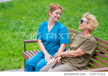 A nurse and an elderly blind woman are sitting on a bench in the park. A nurse and an elderly blind woman are sitting on a bench in the park. 118772122