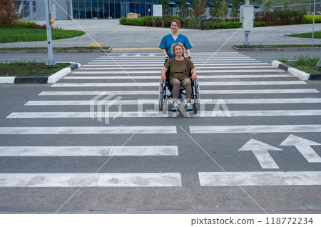 Red-haired nurse pushing an elderly woman in a wheelchair across the road. Red-haired nurse pushing an elderly woman in a wheelchair across the road. 118772234