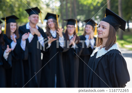 Group of happy students in graduation gowns outdoors. A young girl in the foreground. Group of happy students in graduation gowns outdoors. A young girl in the foreground. 118772265
