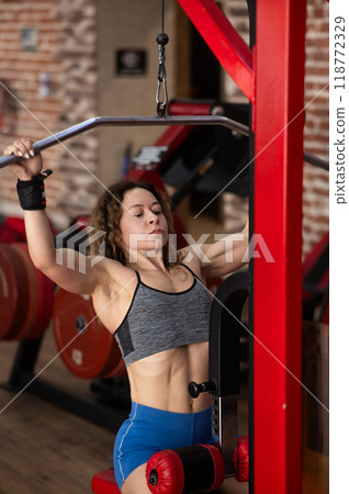 Caucasian forty-year-old woman doing lat pull-downs in the gym. Vertical photo.  118772329