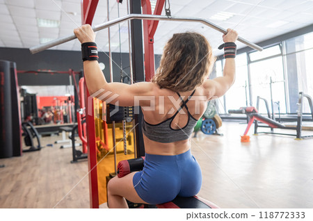 Caucasian forty-year-old woman doing lat pull-downs in the gym. Caucasian forty-year-old woman doing lat pull-downs in the gym. 118772333