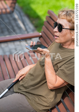 An elderly blind woman sits on a bench in the park and talks on a smartphone. Vertical photo. 118772399