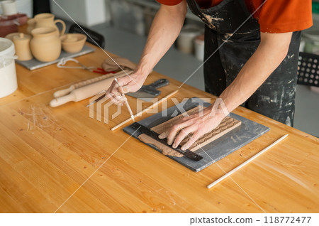 Close-up of a potter's hands cutting off a piece of rolled clay with patterns.  118772477