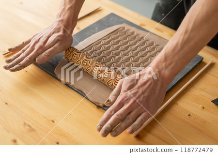 Close-up of a potter's hands rolling out clay using a rolling pin with patterns.  118772479