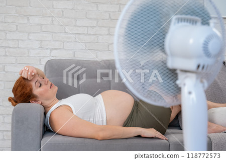 Pregnant woman suffers from heat and lies on the sofa near the electric fan.  118772573