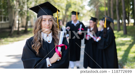 Group of happy students in graduation gowns outdoors. A young girl with a diploma in her hands in the foreground. 118772699