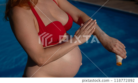 Close-up of the belly of a pregnant woman sunbathing in a red bikini. Expectant mother applying sunscreen to her hands.  118772892