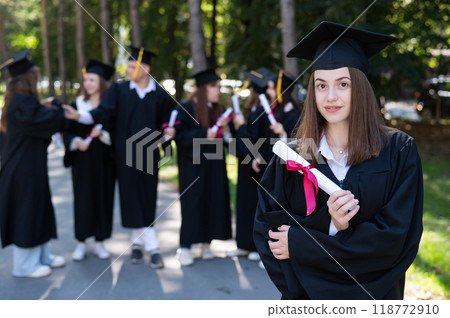 Group of happy students in graduation gowns outdoors. A young girl boasts of her diploma. Group of happy students in graduation gowns outdoors. A young girl boasts of her diploma. 118772910