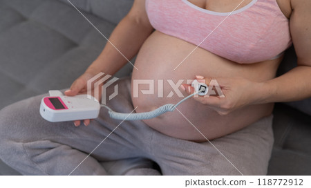 A pregnant woman uses a home doppler while sitting on the couch. Close-up of the abdomen. A pregnant woman uses a home doppler while sitting on the couch. Close-up of the abdomen. 118772912