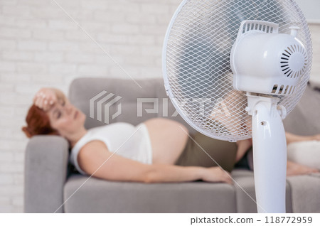 Pregnant woman suffers from heat and lies on the sofa near the electric fan.  118772959