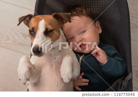 A dog and a cute three-month-old boy dressed in a blue overalls are sitting together in a baby lounger. A dog and a cute three-month-old boy dressed in a blue overalls are sitting together in a baby lounger. 118773048