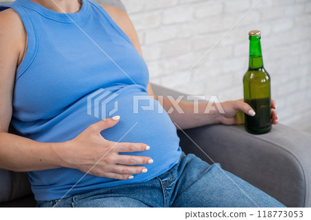 Faceless pregnant woman drinks beer while sitting on the sofa. Faceless pregnant woman drinks beer while sitting on the sofa. 118773053