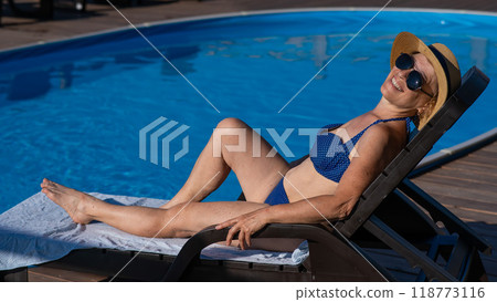 An old woman in a hat and sunglasses is sunbathing on a sun lounger by the pool. Vacation in retirement. An old woman in a hat and sunglasses is sunbathing on a sun lounger by the pool. Vacation in retirement. 118773116