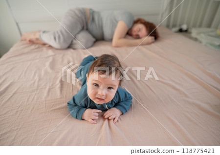 A three-month-old boy lies on his stomach on the bed and his mother sleeps behind him. Postpartum depression. 118775421