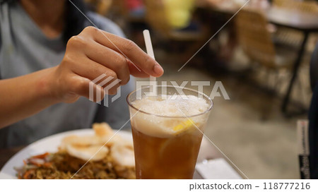 A hand holding straw and drinking a glass of ice lemon tea on the table. A hand holding straw and drinking a glass of ice lemon tea on the table. 118777216