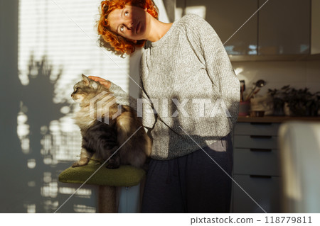 portrait red-haired curly young woman with pet gray fluffy cat in arms, lit by bright sun 118779811