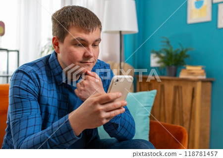 Young Caucasian man text messaging on smartphone sitting on sofa watching online movie at home room 118781457