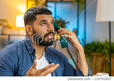 Tired Indian man freelancer talking on retro wired telephone annoyed boring conversation at office 118781607