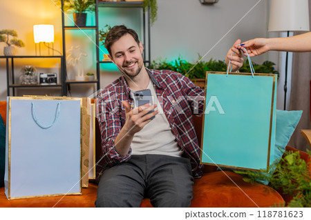Man using smartphone and receiving shopping bags online orders from courier sitting on sofa at home 118781623