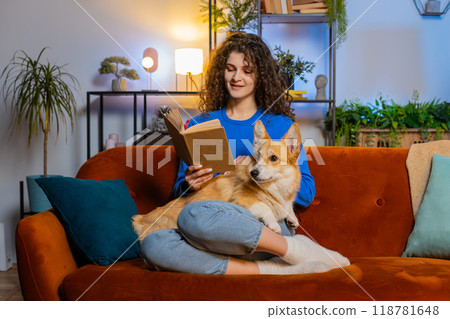 Happy woman reading book while sitting beside cute corgi pet dog on sofa in living room at home 118781648