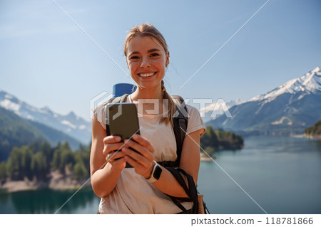 A smiling hiker with a smartphone stands by a beautiful mountain lake, capturing natures beauty A smiling hiker with a smartphone stands by a beautiful mountain lake, capturing natures beauty 118781866
