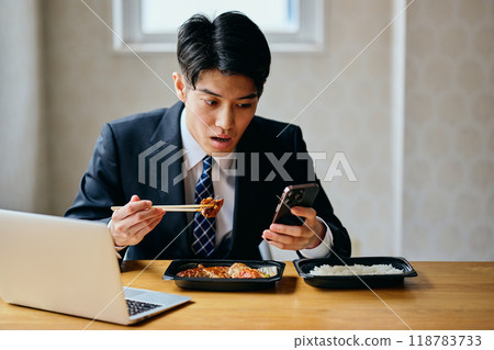 一名20多歲的年輕人一邊吃便當一邊操作智慧型手機 一名20多歲的年輕人一邊吃便當一邊操作智慧型手機 118783733