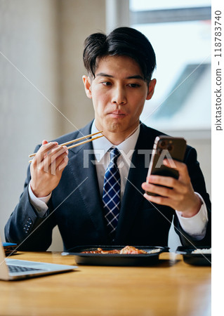 一名20多歲的年輕人一邊吃便當一邊操作智慧型手機 一名20多歲的年輕人一邊吃便當一邊操作智慧型手機 118783740