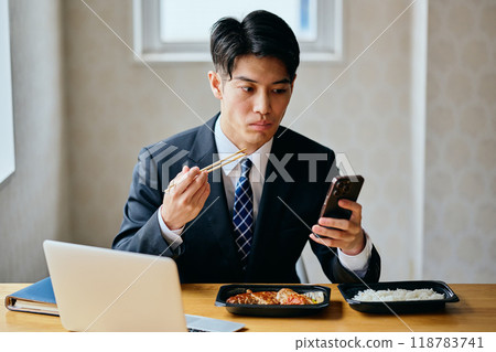 一名20多歲的年輕人一邊吃便當一邊操作智慧型手機 一名20多歲的年輕人一邊吃便當一邊操作智慧型手機 118783741