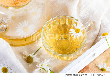 Close up glass cup full of chamomile tea, glass teapot, daisies on white napkin on wooden tray. Close up glass cup full of chamomile tea, glass teapot, daisies on white napkin on wooden tray. 118785239