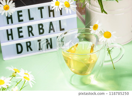 Herbal medicine. Healthy chamomile tea in glass cup, flowers, inscription by black letters on green. Herbal medicine. Healthy chamomile tea in glass cup, flowers, inscription by black letters on green. 118785250