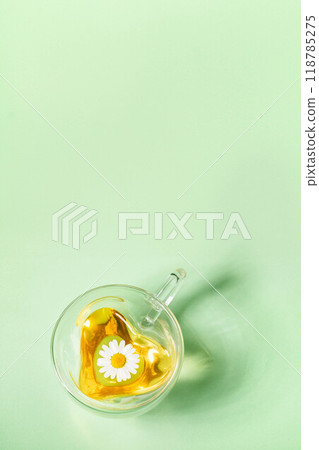 Heart glass cup with chamomile tea with one flower on light green vertical background. Copy space. 118785275
