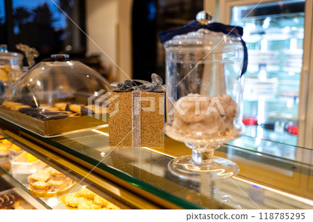 Decorative golden shiny box tied with silver ribbon surrounded by desserts. Christmas or New Year decoration in a cafe. 118785295