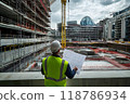 Engineer reviewing a large civil engineering project blueprint at a construction site. The architect wears overalls, a vest and a helmet. Engineer working at site of a large building project. 118786934