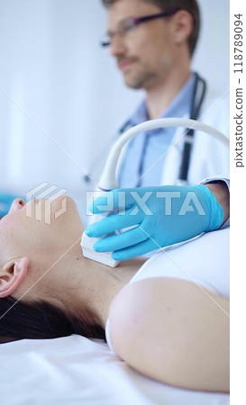 Doctor man wearing blue protective gloves is using ultrasound machine to examine neck of female patient in clinic. Medicine, thyroid gland examination 118789094