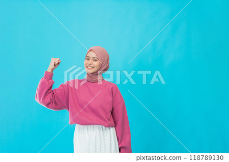 A Confident Young Woman is Celebrating Her Success with a Joyful Fist Bump Against a Blue Background A Confident Young Woman is Celebrating Her Success with a Joyful Fist Bump Against a Blue Background 118789130