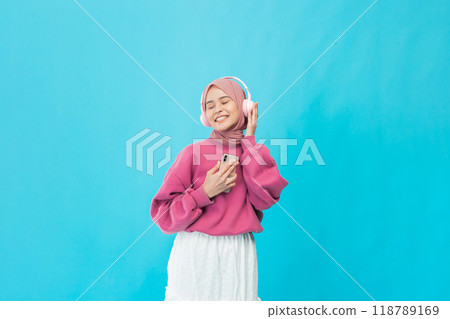 A carefree young woman enjoys music in a stylish pink sweater against a vibrant blue background A carefree young woman enjoys music in a stylish pink sweater against a vibrant blue background 118789169