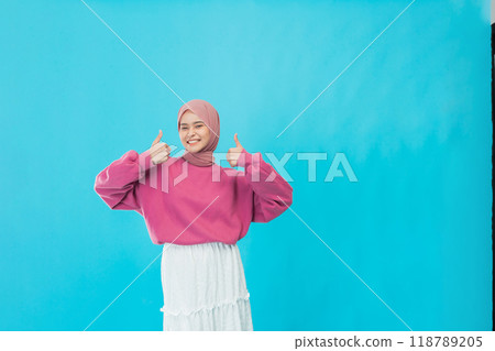 A Cheerful Young Woman Wearing a Pink Sweater is Giving a Thumbs Up Against a Bright Blue Background 118789205
