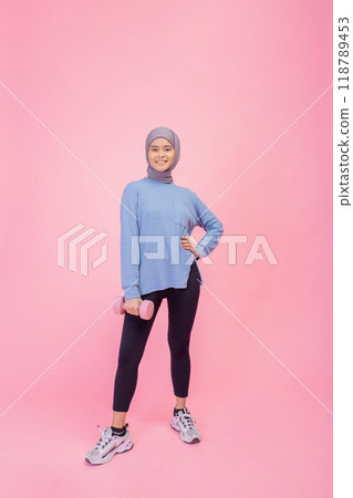 An athletic woman holds a dumbbell in stylish activewear against a vibrant pink background 118789453