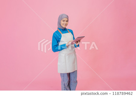 A young woman in a stylish apron holds a tablet, standing confidently against a bright pink background 118789462