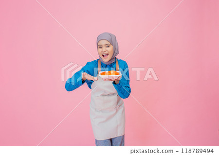 A cheerful female chef presents a delicious dish against a pink background, showcasing her happiness 118789494