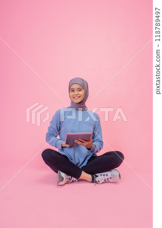 A Young Woman with a Hijab is Sitting Relaxed while Using a Tablet on a Pink Background A Young Woman with a Hijab is Sitting Relaxed while Using a Tablet on a Pink Background 118789497