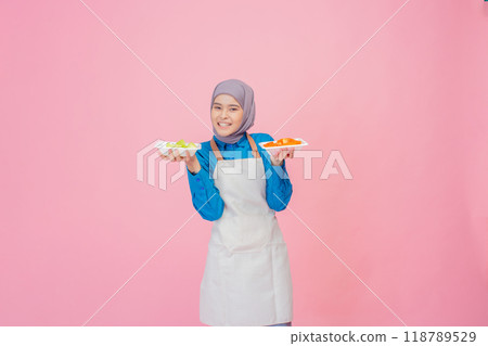 A Young Woman in a Stylish Apron Holding Plates of Fresh, Colorful Vegetables Against a Pink Background 118789529