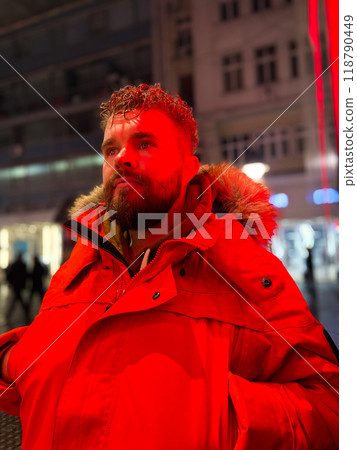 Man walking in a bustling nighttime city street, wearing a bright orange parka. Red neon light. Serene contemplation against the backdrop of lively city lights at night. Urban life concept 118790449