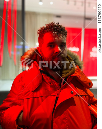 Man walking in a bustling nighttime city street, wearing a bright orange parka. Red neon light. Serene contemplation against the backdrop of lively city lights at night. Urban life concept Man walking in a bustling nighttime city street, wearing a bright orange parka. Red neon light. Serene contemplation against the backdrop of lively city lights at night. Urban life concept 118790450